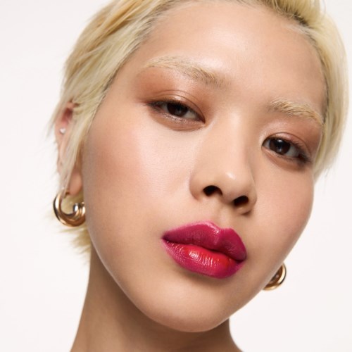 Nahaufnahme einer Frau mit kurzem blonden Haar, mutiger Make-up, rosa Lippenstift, Ohrringen und selbstbewusstem Ausdruck.