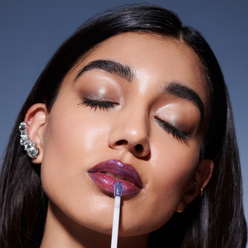 A woman with closed eyes, shiny makeup, and purple lipstick applying gloss with a stick.