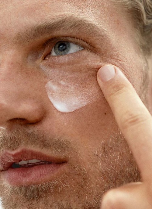 Ein Mann trägt Creme unter seinem Auge auf und berührt die Stelle mit seinem Finger. Nahaufnahme der Hautpflege.