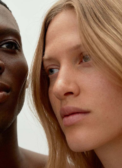 Zwei Frauen stehen nah beieinander, eine mit dunkler Haut, die andere mit heller Haut und blonden Haaren, beide sehen ernst aus.