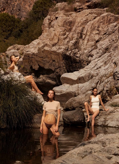 Drei Frauen in Badebekleidung sitzen und stehen auf Felsen bei einem kleinen Wasserlauf, umgeben von felsigem Gelände und spärlicher Vegetation.