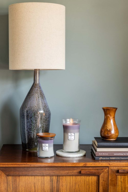 Een hoge lamp, twee kaarsen, een klein houten vaasje en boeken staan op een houten tafel tegen een effen muur.