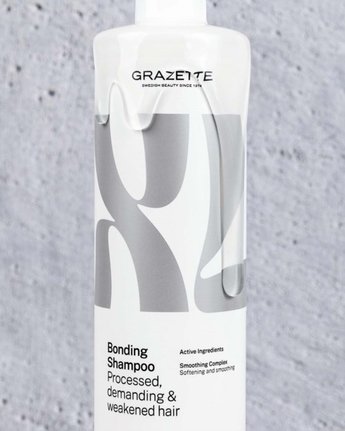 Een witte flesje van Grazeite Bonding Shampoo met grijs abstract ontwerp op het label, tegen een grijze textuurrijke achtergrond.