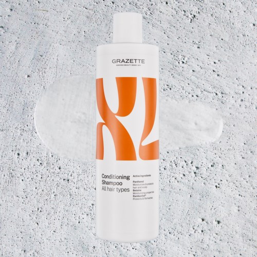 En vit flaska med GRAZETTE Balsam Shampoo på en texturerad yta med en schamposkvätt bakom.