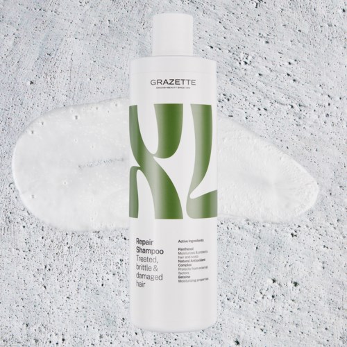 Een witte fles GRAZETTE Repair Shampoo met groen ontwerp, geplaatst op een witte textuurrijke ondergrond met crèmige strepen.