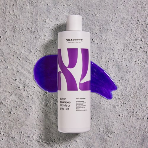 Eine weiße Flasche mit der Aufschrift 'GRAZETTE Silver Shampoo' und lila Design liegt auf einer grauen Oberfläche mit lila Farbe.