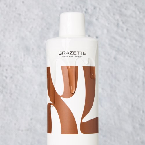 Eine weiße Flasche mit braunen und weißen Texten, vor einem strukturierten grauen Hintergrund.