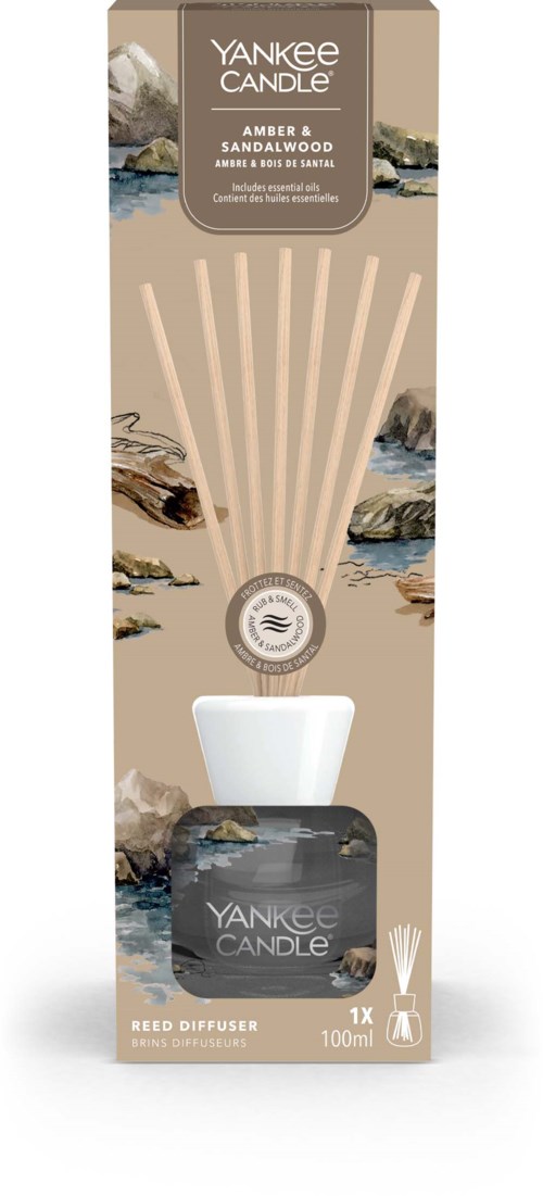 Een doos met geurverspreiders, beige en zwart, met label "Yankee Candle Amber Sandalwood," met stokjes erin.