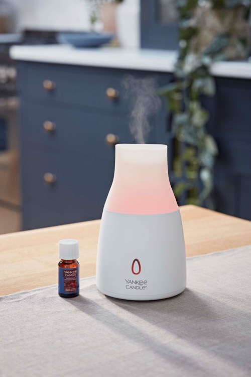 En vit diffuser med rosa sken avger ånga bredvid en liten äkt essential olja på ett trädgårdsbord.