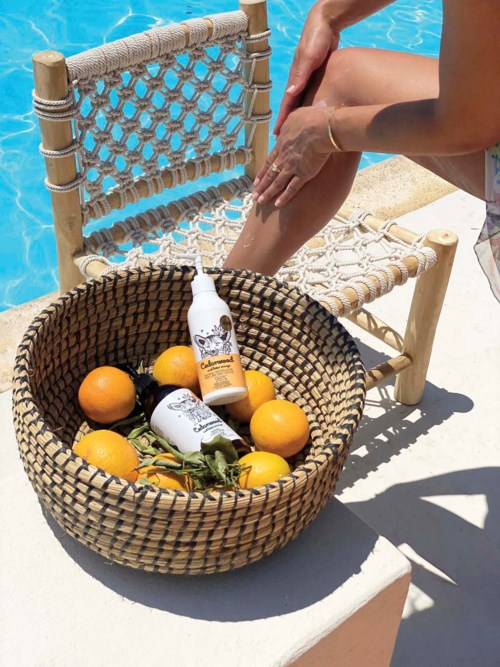Ein Korb mit Orangen und Hautpflegeprodukten neben einem kleinen Stuhl am Pool. Eine Person cremt sich die Beine ein.