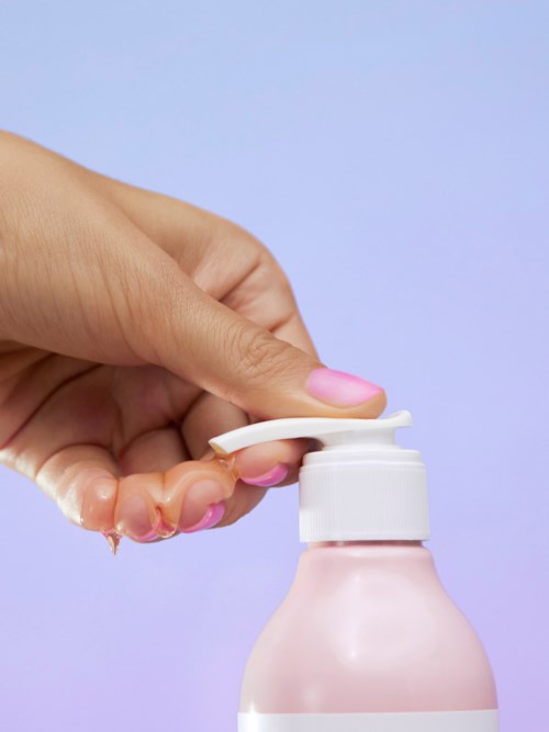 Eine Hand drückt auf eine Pumpe an einer rosa Flasche, wahrscheinlich für Seife oder Lotion, vor einem helllila Hintergrund.