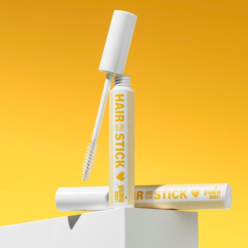A hair wax stick with a white cap and yellow packaging, placed on a white surface against a yellow background.