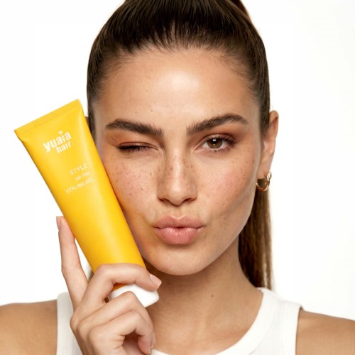 A woman with a tied-back hairstyle making a face and holding a yellow beauty product tube.