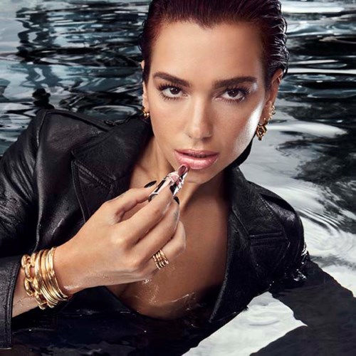 A woman in a black leather jacket is partially submerged in water, applying lipstick, with jewelry and a serious expression.