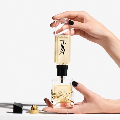 Iemand houdt een glazen fles met parfum en giet het in een kleinere container. De achtergrond is eenvoudig en wit.