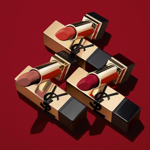 Drei Lippenstifte in Pink-, Pfirsich- und Rottönen, in Gold- und Schwarzdosen auf rotem Hintergrund.