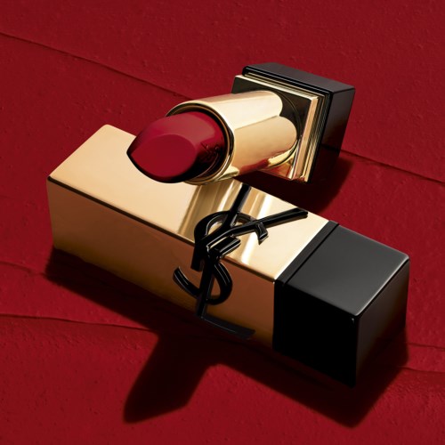 A YSL lipstick with red color and gold packaging lies on a red background. The shadow of the lipstick is visible.