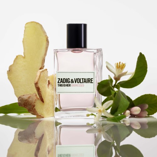 Eine Parfümflasche mit dem Label Zadig & Voltaire, umgeben von Ingwerscheiben, weißen Blumen und grünen Blättern.