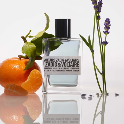 Ein Parfümflasche mit Orange, Zitrone und Lavendel drumherum auf einer reflektierenden Oberfläche.