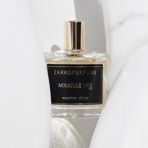 Hajuvesilasi, jossa musta etiketti, kultainen kansi ja kirkas lasi,valkoisella pinnalla.