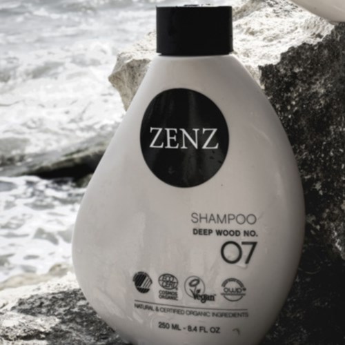 A white shampoo bottle named ZENZ on rocks near water. It has black and white labels with organic and vegan symbols.