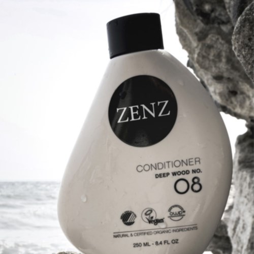 A white bottle labeled ZENZ conditioner, with a black cap, is placed outdoors near rocks and the sea.