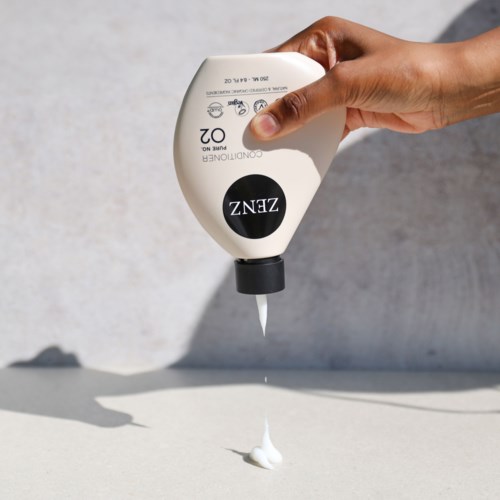 A hand pouring white liquid from a tube onto a flat surface. The tube is labeled ZENZ and appears to be shampoo.