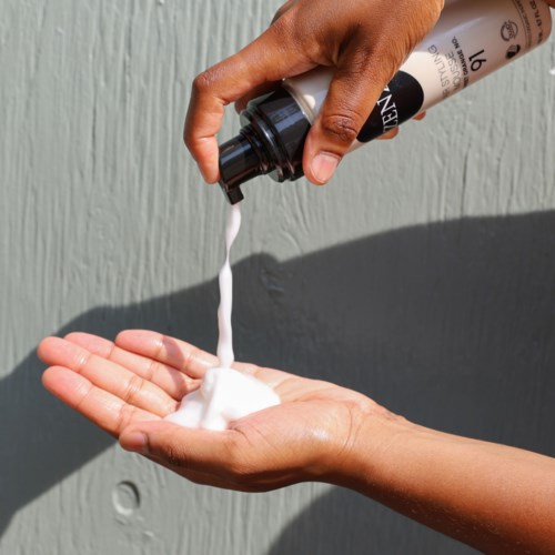 A hand holds a soap bottle, pouring soap into an open palm against a light background.