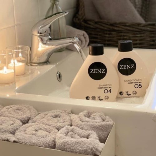 Two bottles of ZENZ shampoo and conditioner sit on a bathroom sink, with rolled towels and candles nearby.