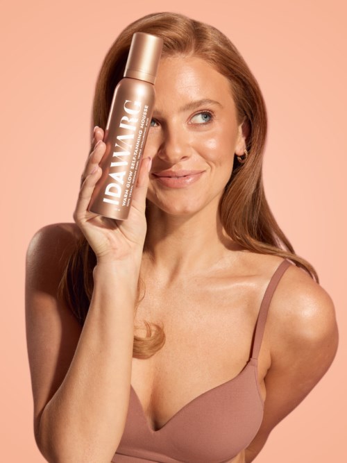 A woman with long red hair holds a bottle of skincare product near her face, smiling softly.