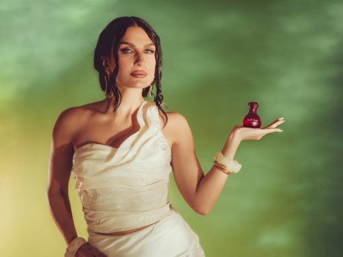 A woman in white dress holds a small red perfume bottle on her open palm against a green background.