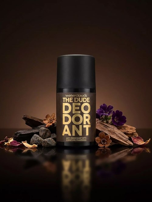 The image shows a black deodorant can with gold text, surrounded by rocks, wood pieces, and purple flowers on a dark background.