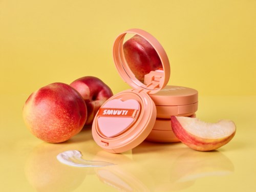 En necessär med spegel omgiven av äpplen och peach-färgade produktbehållare på en gul yta.