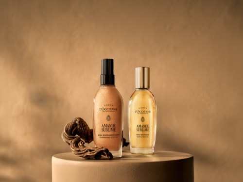 Två flaskor av L'Occitane Amande Sublime produkter med en hårlock nära flaskorna på en rund plattform.