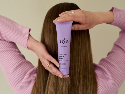 En person med langt brunt hår holder en lilla flaske med hårkrem bak hodet, iført en rosa genser.