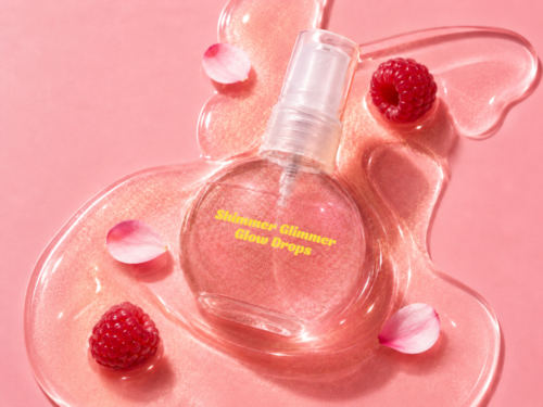 En klart sprayflaska märkt Shimmer Gliner Glow Drops omgiven av hallon och rosa blomblad på rosa bakgrund.