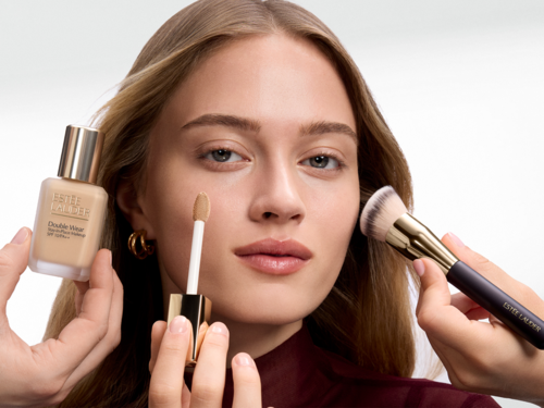En kvinna med långt hår får makeup applicerad; en flaska foundation, ett concealerstift och en makeupborste syns.