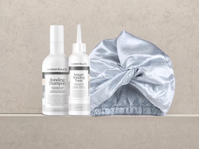 Två vita flaskor med hårvårdsprodukter, en märkt Bombing Shampoo och den andra Instant Bonding Tonic, bredvid en silverfärgad satinmössa.