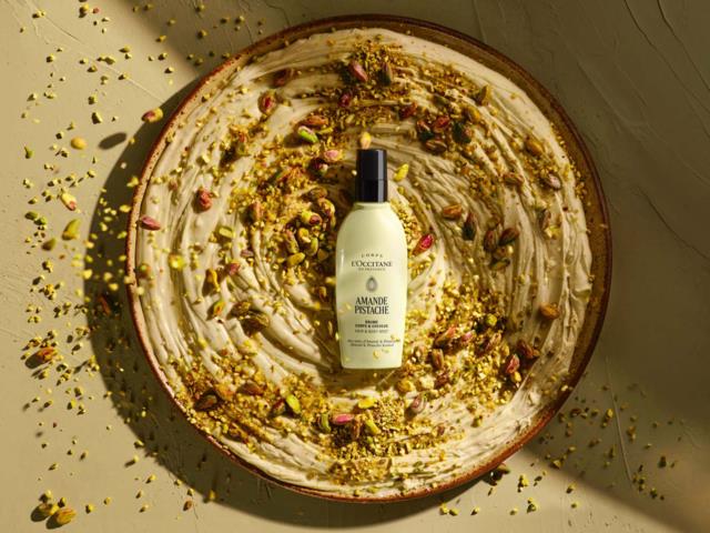 En flaska L'Occitane almond shower oil placerad i en dekorativ skål med krossade pistager runt om. Ljust ljus.