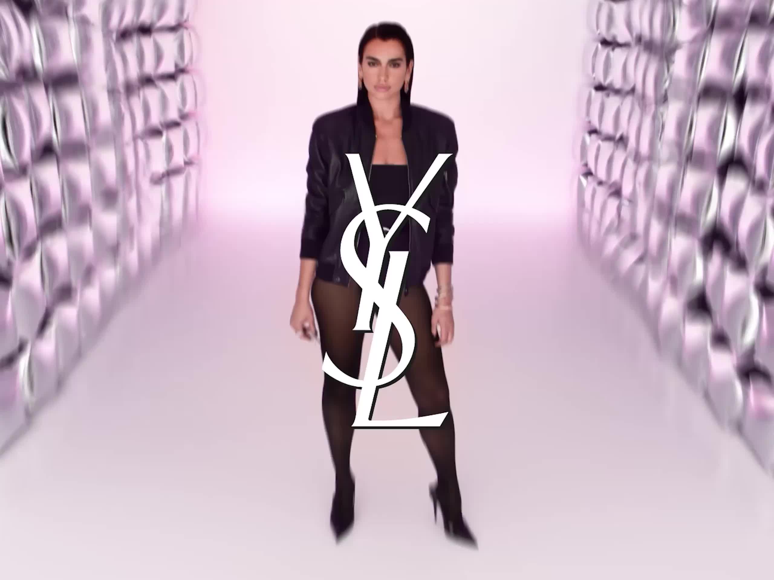 A woman in black standing with a large YSL logo on her outfit, shelves with shoes on both sides, pink background.